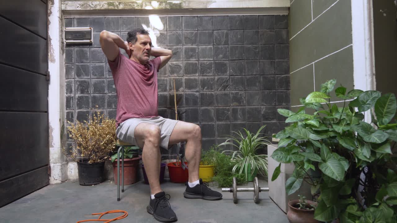 A focused, active man sits on a chair in his indoor patio, performing a controlled, modified bicycle crunch to engage his core and abdominal muscles