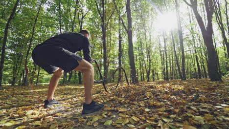 strong man doing waves with ropes during workout. backside view of a young athlete exercising with black ropes among trees in the forest. slow motion.