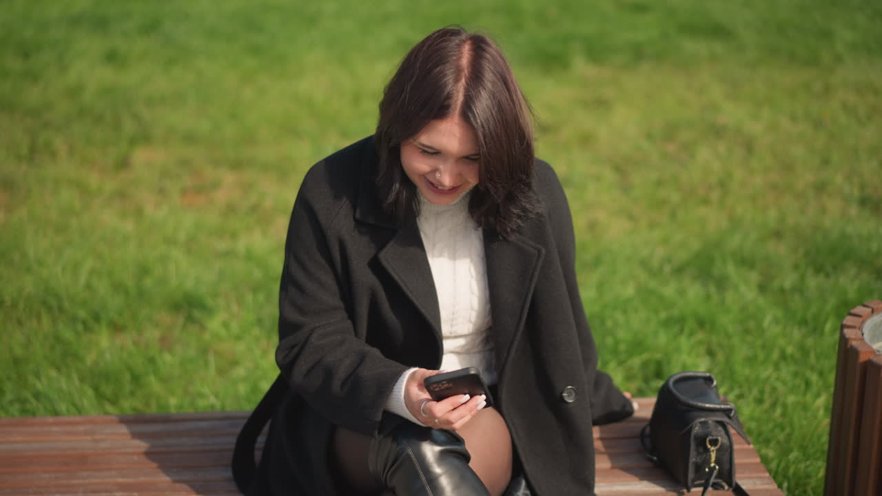 공원에서 앉아서 스마트폰에 집중하는 미소 짓는 여인, 검은 코트와  스웨터를 입고, 다리를 고, 울창한 초록색으로 둘러싸여