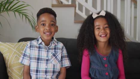 african american siblings siting on sofa, looking at camera and smiling