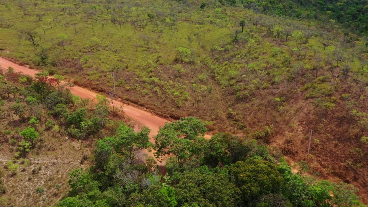 En drone følger en grå 4x4 pickup og glider derefter ind i et push-in mod den blå Tocantins-velkomstportal, der spænder over en lige, rød jordvej i Cerrado, Brasilien.