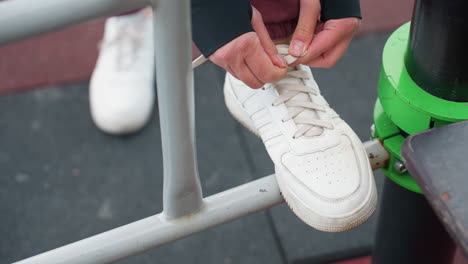 high angle view of woman tying sneaker laces on iron workout bar with foot perched on metal rail before urban outdoor fitness routine near modern apartment complex on rubber mat surface