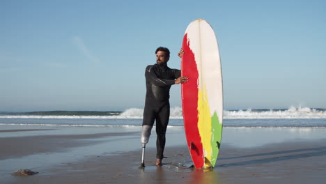 long shot of a male surfer in wetsuit with artificial leg standing on the beach while leaning on the surfboard