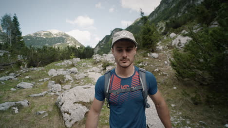 camera tracking hiker infront on his path on the mountain, wearing a grey hat, walking with hiking poles, sunny summer day with some clouds on the sky