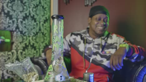 a black man is stoned after taking a hit from a marijuana ice filled water bong the cannabis smoking device is sitting on the table with a bag of weed