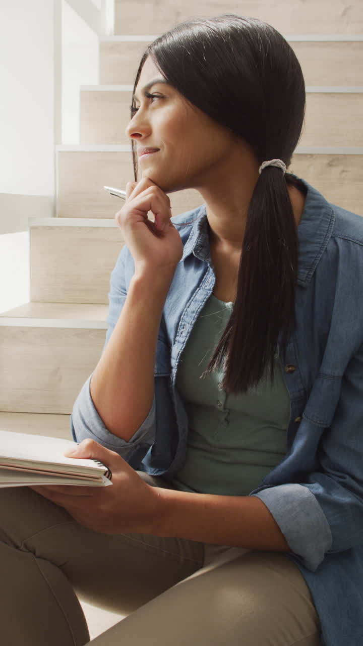 video vertical de una mujer biracial pensativa sentada en las escaleras y tomando notas