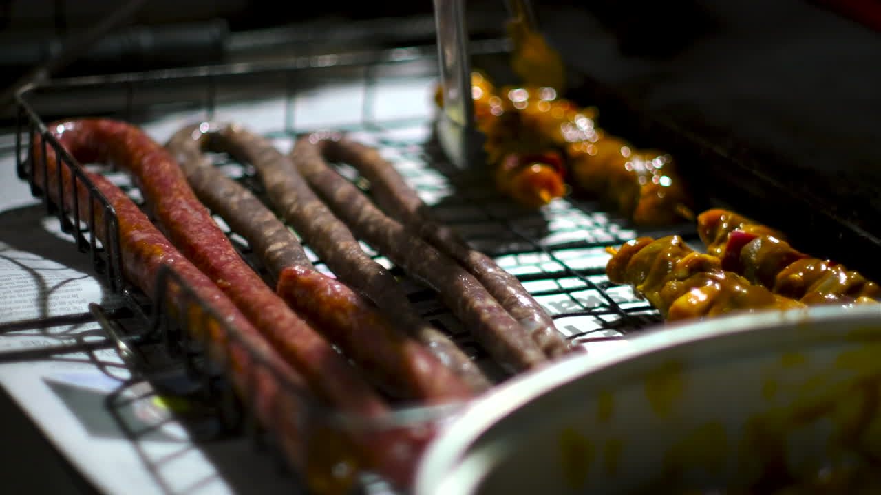 Using tongs to place marinated lamb skewers onto grid near sausages for barbecue