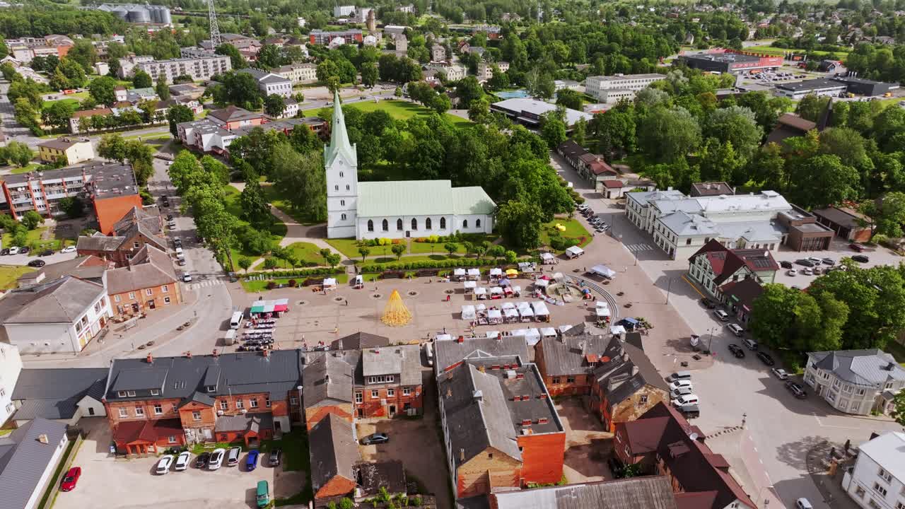 Luftaufnahme der Kleinstadt Dobele mit Kirche und Marktplatz unter bewölktem Himmel.
