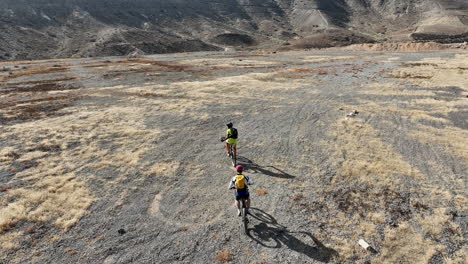 Fantástica-Toma-Aérea-De-Dos-Hombres-Con-Sus-Bicicletas-De-Montaña-Que-Recorren-La-Carretera-A-Alta-Velocidad-En-Un-Paisaje-Desértico-Con-Montañas