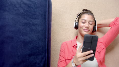 smiling biracial teenage girl in headphones lying on floor using smartphone, copy space, slow motion