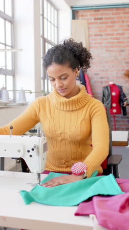 diverse female fashion designer sewing fabric with sewing machine in studio, slow motion, vertical