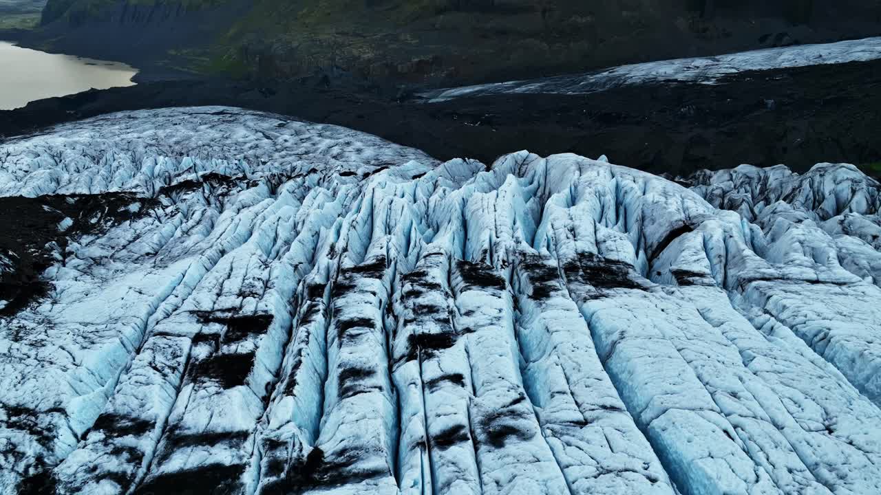 Droneoverflyvning af en islandsk gletsjer med dybe sprækker og mørke vulkanske askebånd, der viser et robust, frossent terræn under et dystert, overskyet lys