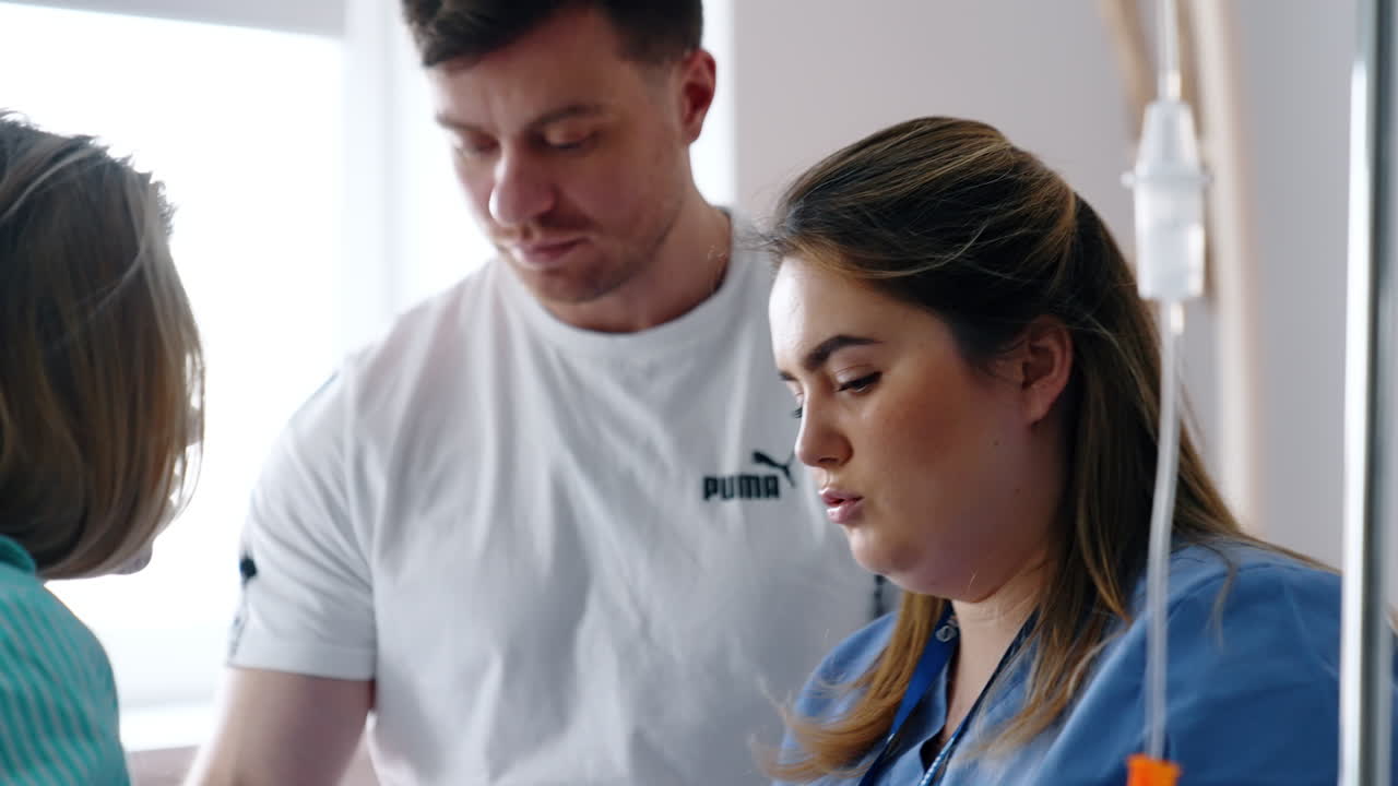 Female medic talks to a couple expecting a child. Nurse explains something to a pregnant woman sitting on the bed her back to camera. Male at backdrop.