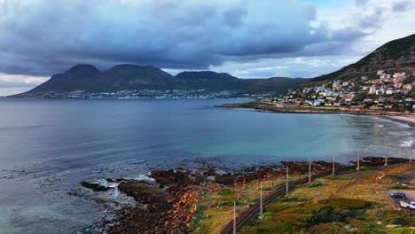 South-African-sunrise-sunset-Simon's-Town-drone-aerial-cinematic-Fish-Hoek-rugged-reef-coast-small-fishing-town-marina-city-stunning-summer-spring-deep-aqua-blue-ocean-train-tracks-backward-movement