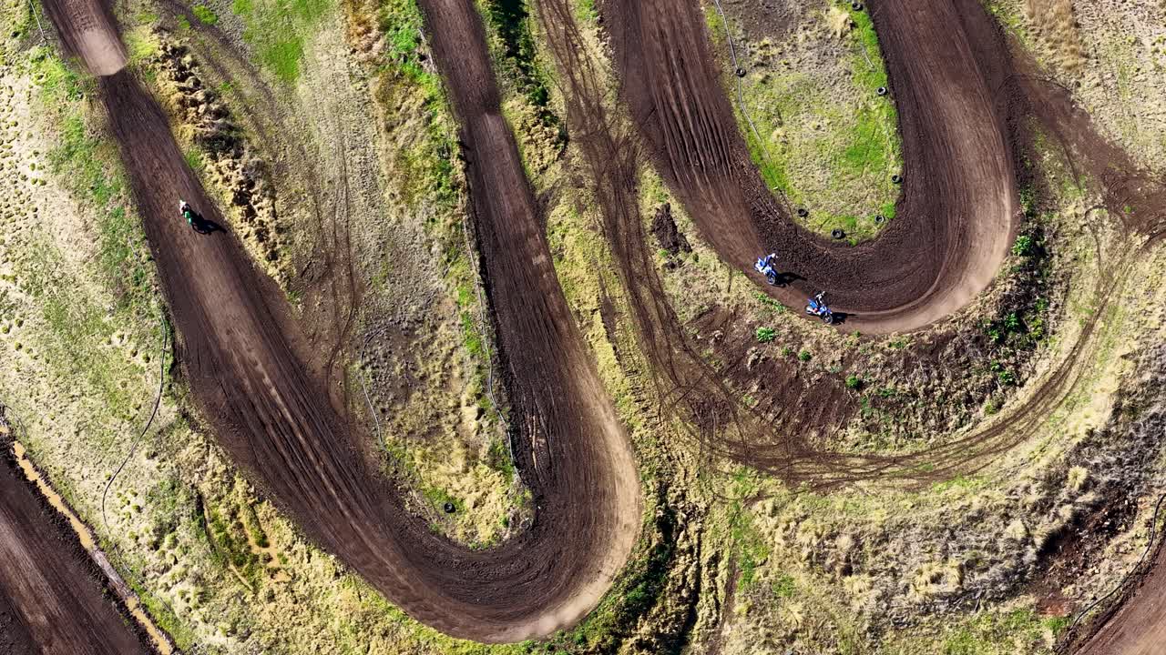 Motorbikes speed around a winding dirt track curve, captured from above in bright daylight