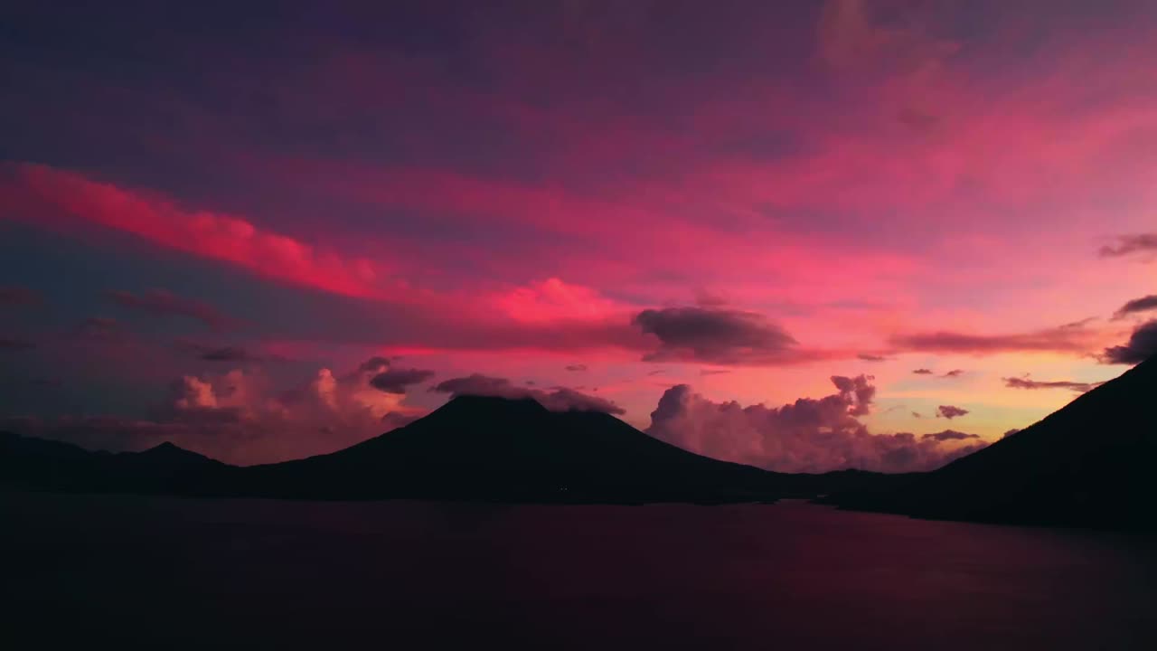 Bredt luftfoto af søen Atitlan i tusmørke med intense magentafarvede skyer, mørke vulkanske silhuetter og stille vand
