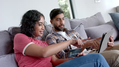 Feliz-Pareja-Birracial-Sentada-En-El-Sofá-Y-Usando-Una-Tableta-Para-Comprar-En-Línea-En-Casa,-En-Cámara-Lenta