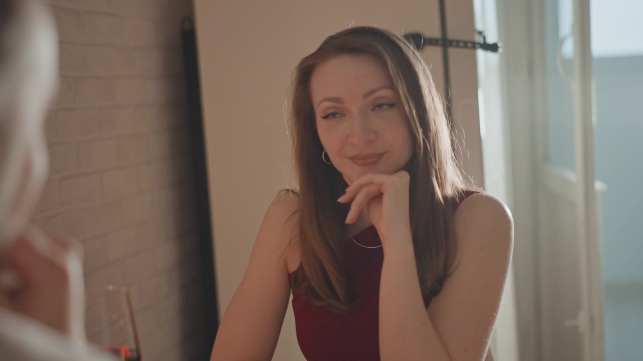 Young woman seated at table touches chin, tilts head, and smiles with eyes softly closed during conversation beside blurred man in foreground, inside sunlit room with warm natural light