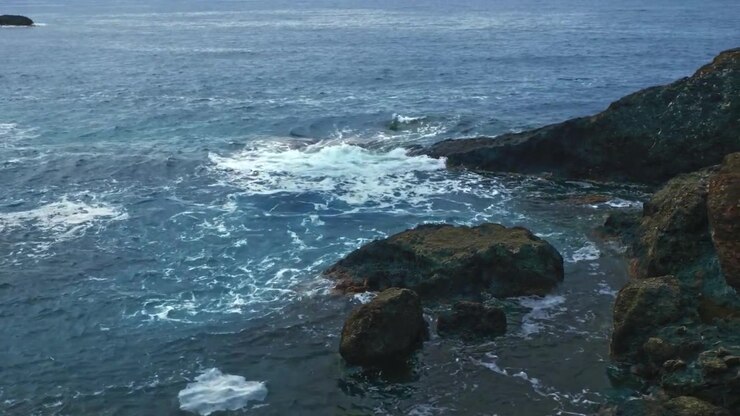Le onde si infrangono contro scure rocce frastagliate lungo la frastagliata costa del Mar Ligure vicino a Riomaggiore nelle Cinque Terre, Italia. Schiuma bianca si diffonde sull'acqua.