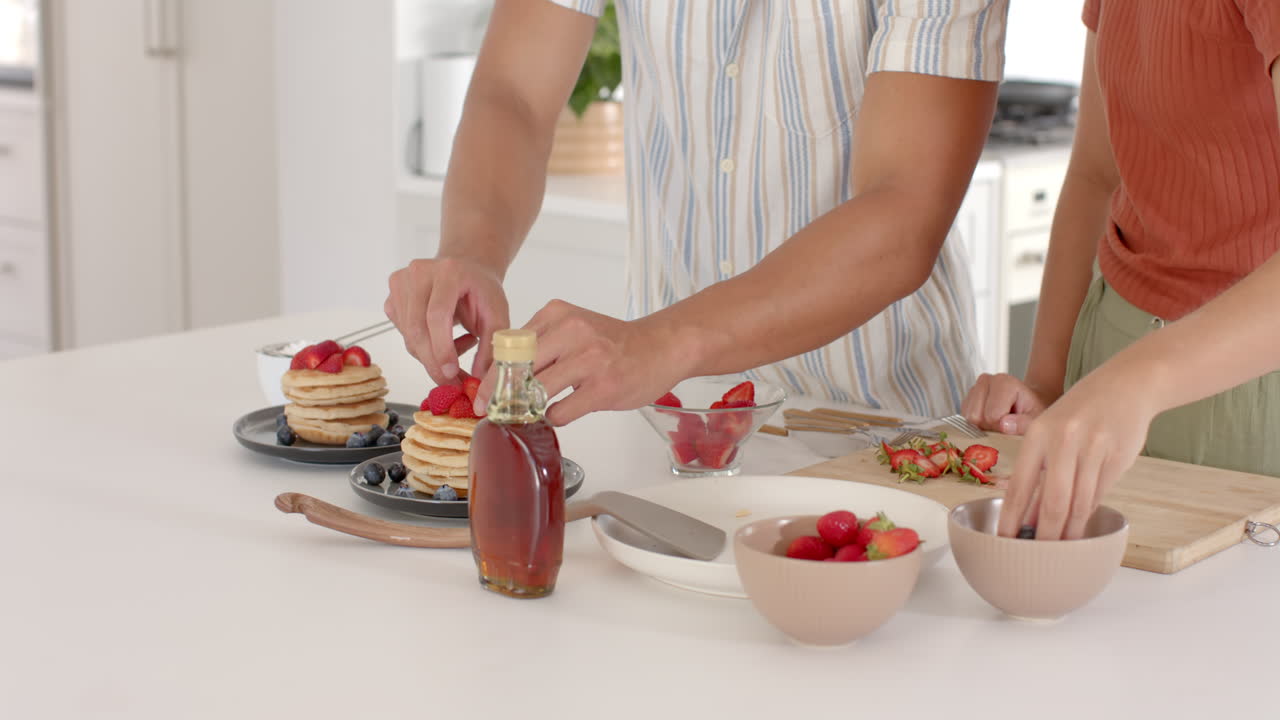 Diverse couple preparing breakfast with pancakes and fresh strawberries in modern kitchen, at home
