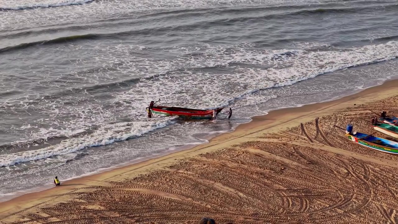 Veduta aerea da drone di una barca da pesca che si avvicina a una spiaggia sabbiosa attraverso dolci onde marine vicino alla costa di Chennai all'alba, catturando l'attività di pesca costiera tradizionale.