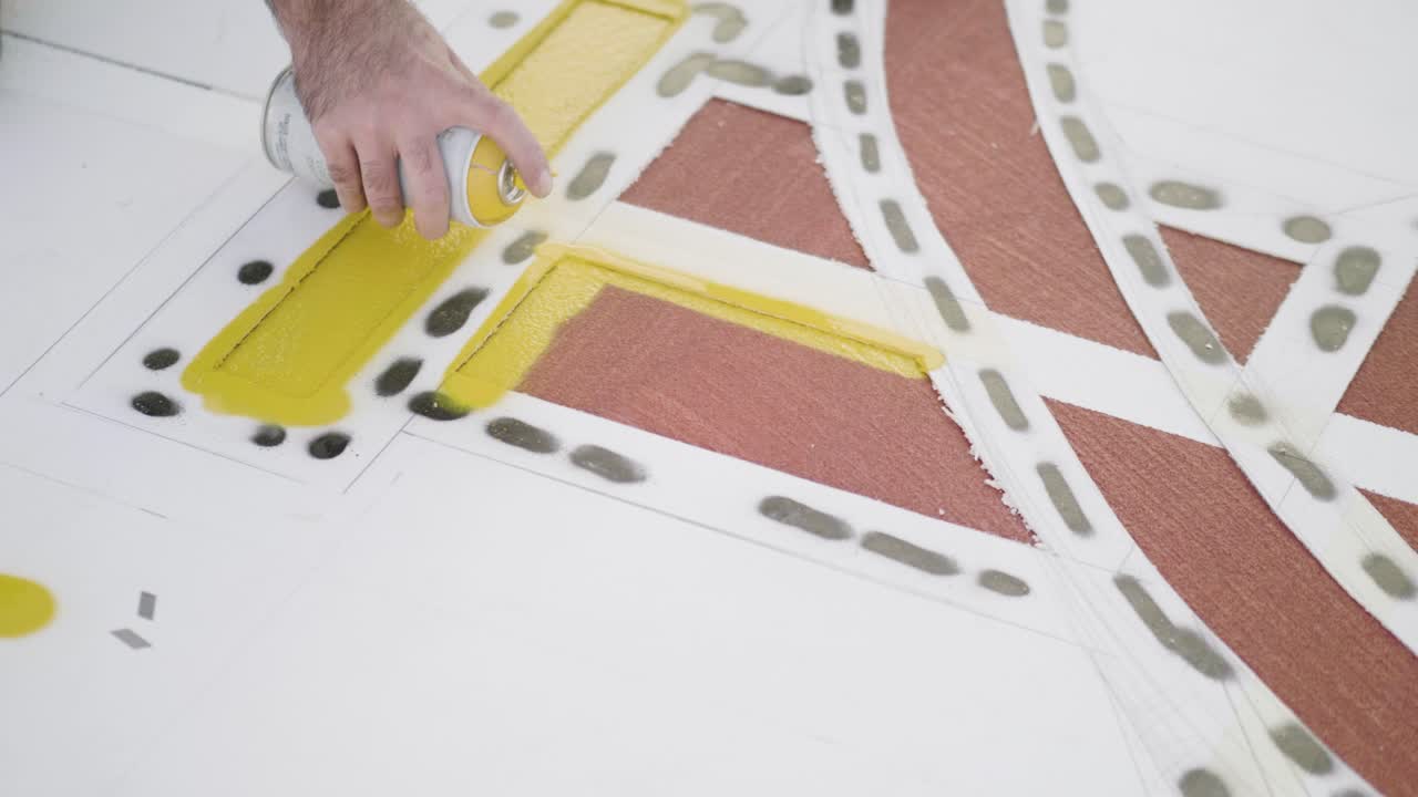Close-up of hand applying yellow spray paint on geometric stencil over white surface, showcasing artistic technique and craft skills, drone static shot, real time