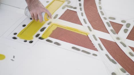close-up of hand applying yellow spray paint on geometric stencil over white surface, showcasing artistic technique and craft skills, drone static shot, real time