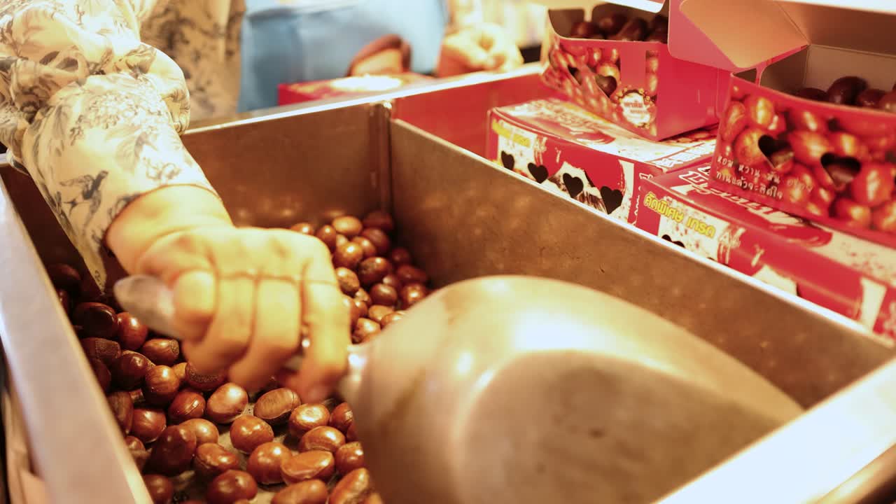Street vendor scoops roasted chestnuts into metal bin under warm lighting, Chinatown, Bangkok, Thailand