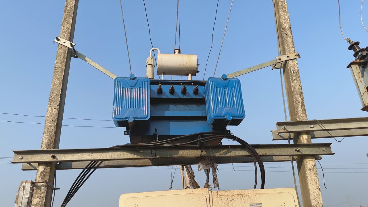 Ripresa con camera orbitante di un trasformatore di distribuzione elettrica montato su palo con alette di raffreddamento blu, connessioni ad alta tensione, isolanti e linee elettriche su un cielo azzurro terso