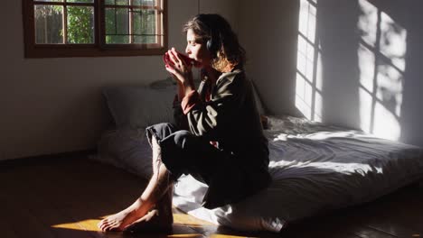 Relaxed-mixed-race-woman-wearing-headphones-sitting-on-bed-drinking-tea-in-sunny-cottage-bedroom