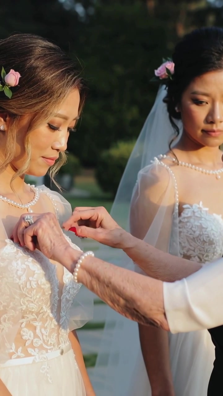 Vertical video: Reaching senior woman fastening bride's pearls and lace dress in garden, preparing