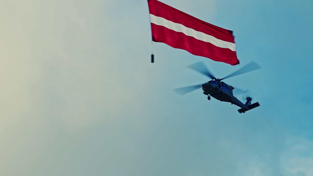 Inquadratura cinematografica dal basso di due elicotteri militari (Sikorsky UH-60 Black Hawk) che eseguono un sorvolo cerimoniale, trasportando la bandiera nazionale rossa-bianca-rossa della Lettonia contro un drammatico cielo blu nuvoloso.