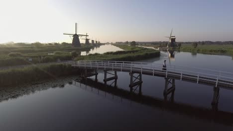 Eine-Drohnenaufnahme-Mit-Stativ,-Die-Ein-Model-Filmt,-Das-Bei-Sonnenaufgang-über-Eine-Brücke-Geht,-Mit-Holländischen-Windmühlen-Im-Hintergrund