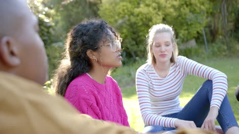 happy diverse group of teenage friends sitting on grass and talking in sunny park, slow motion