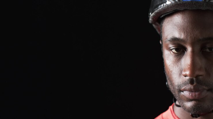 Ciclista afroamericano con casco mirando fijamente sobre un fondo negro, con espacio para texto.
