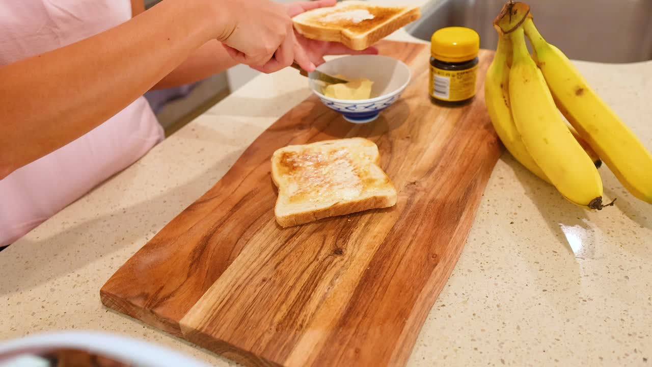 A person spreads butter on toast in a well-lit kitchen, surrounded by bananas and a jar of spread