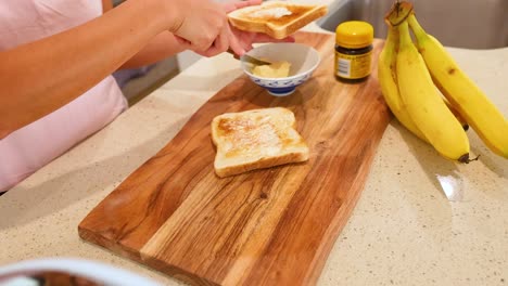 a person spreads butter on toast in a well-lit kitchen, surrounded by bananas and a jar of spread