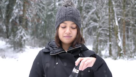 young caucasian woman drinking hot tea from thermo bottle in the park on snowy day