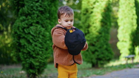 caucasian baby boy holding a big black cap outdoors. toddler puts the cap on, looks at camera and hides face behind it. blurred backdrop.