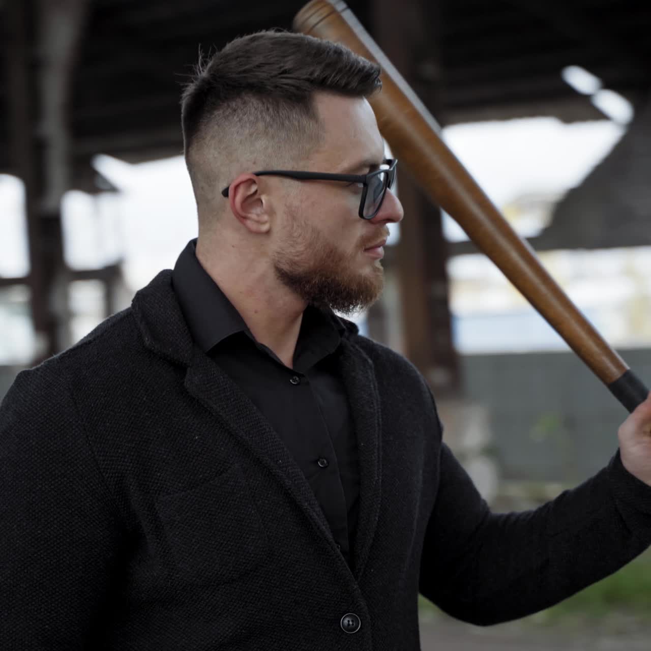 Handsome man in ruined place. Serious bearded man in glasses stands with baseball bat inside the damaged building. Camera moves in around.