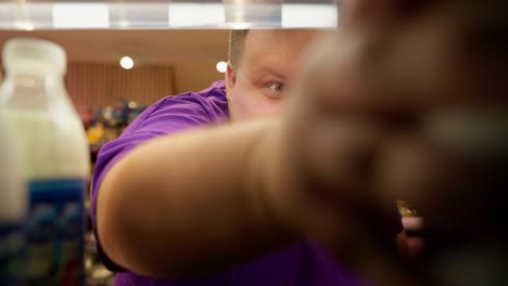Happy overweight man in purple t-shirt raises eyebrow and takes dairy product from shelf in supermarket. Selection of milk in a supermarket