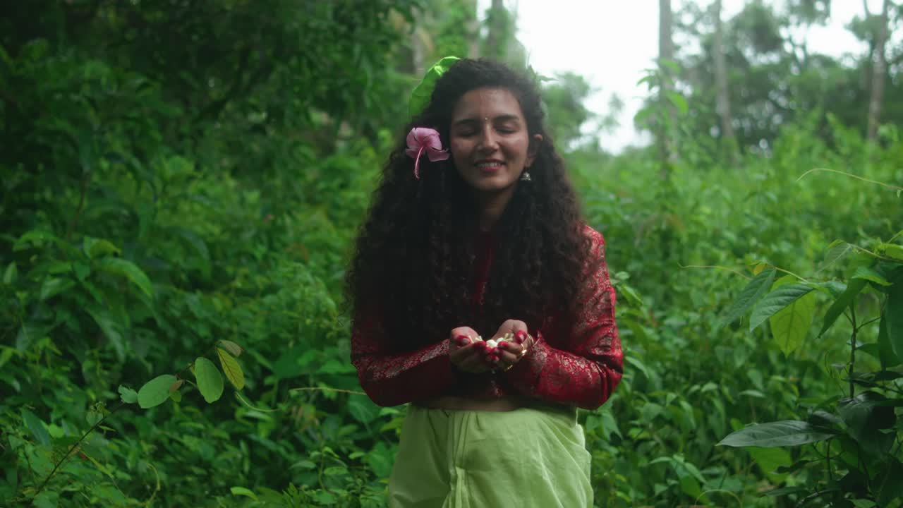 The camera slowly moves toward a smiling woman standing in the jungle, holding white flowers in her cupped hands. She's wearing a green sari, red blouse, and a pink flower in her hair