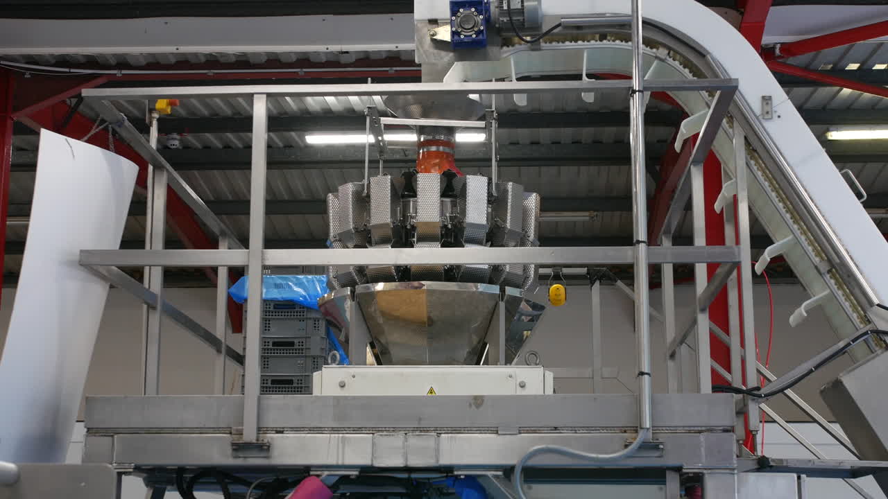 A multihead weigher machine is mounted on a platform in a sweet factory, featuring numerous stainless steel hoppers. A conveyor feeds ingredients into the top for automated weighing and packing