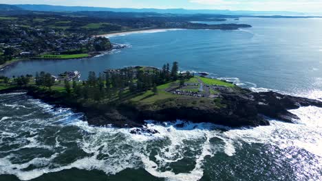 Drone-aerial-landscape-view-of-trees-on-main-town-beaches-bay-rocky-headland-of-Kiama-blowhole-cliffs-and-lighthouse-carpark-South-Coast-Australia-travel-tourism