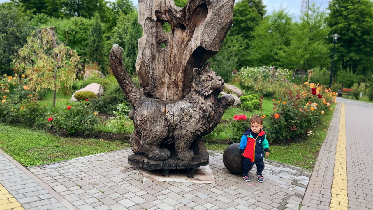 Little kid stands by the wooden sculpture of cat in the park. Cute baby sees the black cat coming up.
