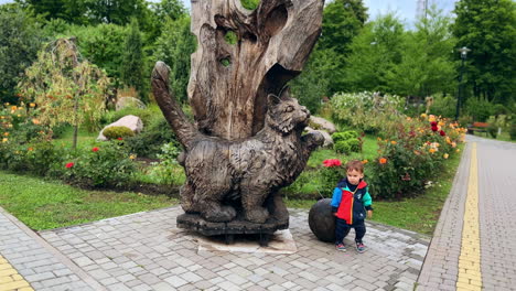 little kid stands by the wooden sculpture of cat in the park. cute baby sees the black cat coming up.