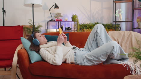 happy young woman in wireless headphones relaxing lying on sofa at home listening favorite music