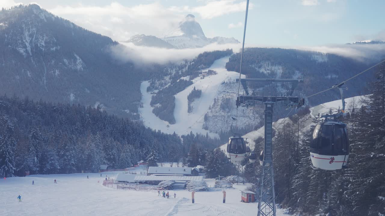 A scenic lift line crosses a snowfield surrounded by pine forest and towering mountain ranges.