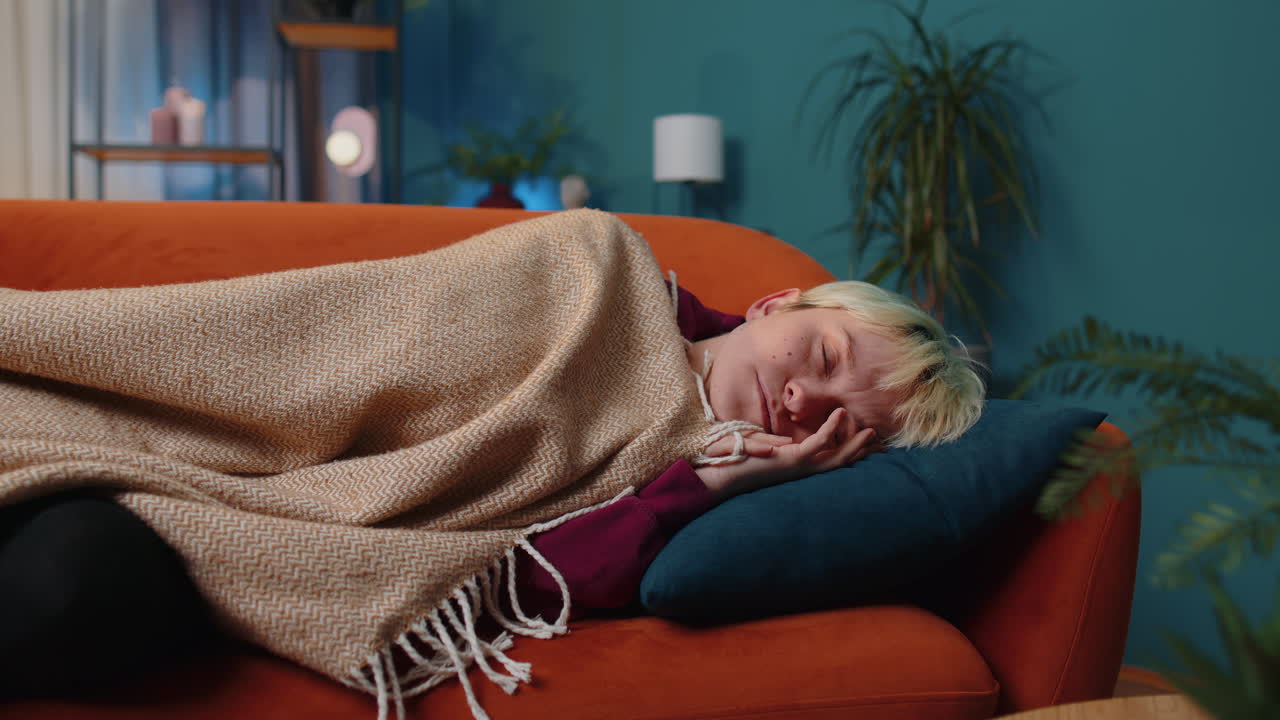 Tired blonde young woman lying down in bed taking a rest at home, napping falling asleep on sofa