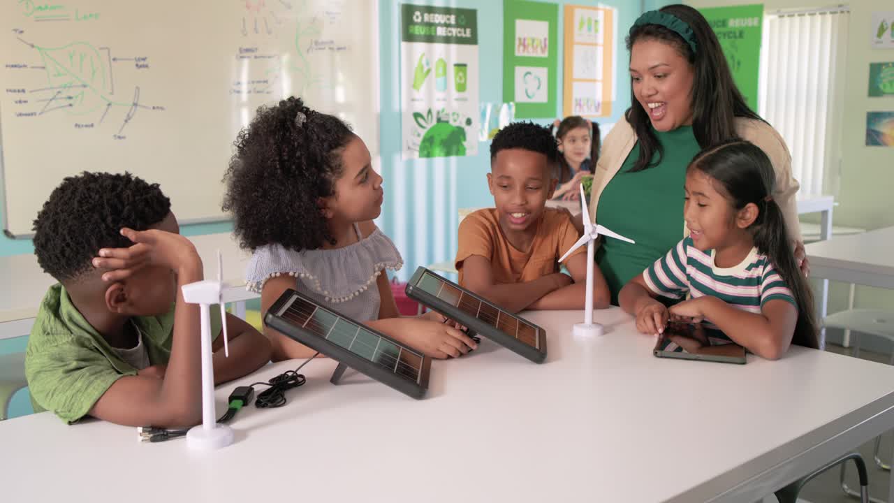 Diverse kids watching girl moving solar panel in STEM class with turbine and tablet teacher nearing
